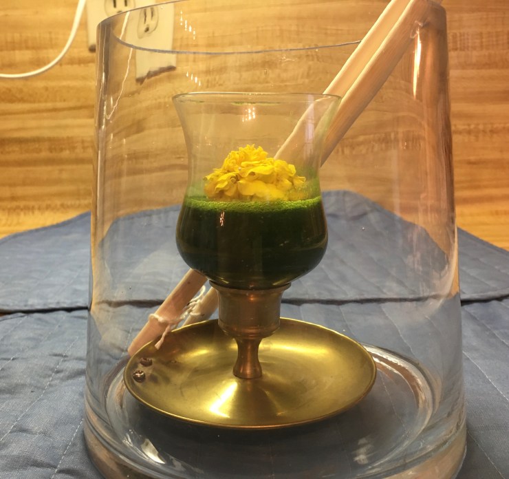 Stinging nettle soup in a glass inside a vase with giant chopsticks in the vase.