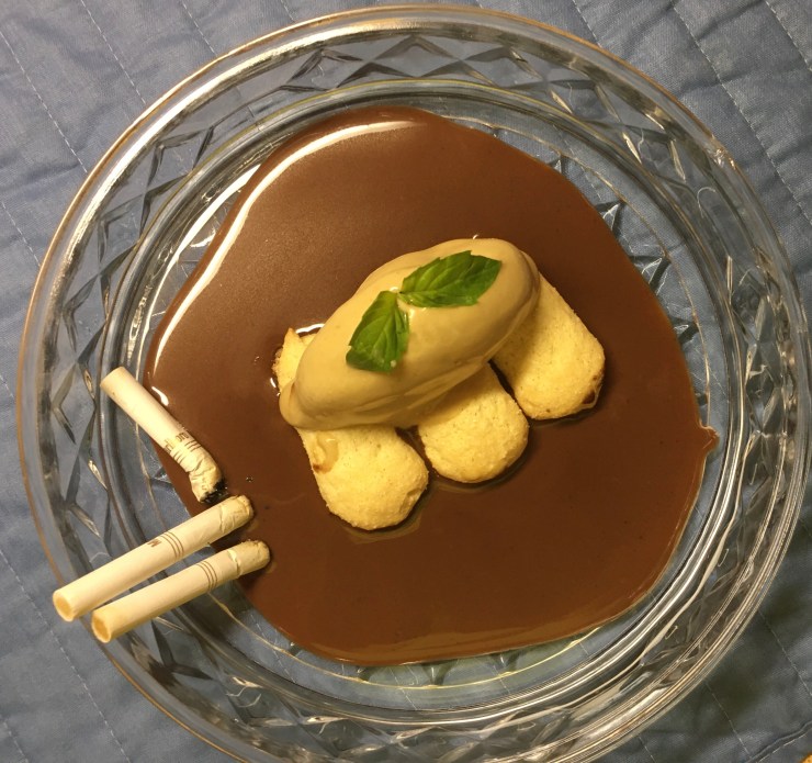 Tobacco ice cream on ladyfingers on chocolate sauce in an ashtray with three old cigarettes on the left side.
