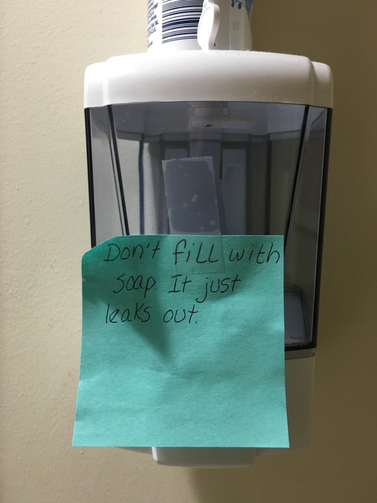 Post it note taped to a soap dispenser that reads, “Don’t fill with soap. It just leaks out.”